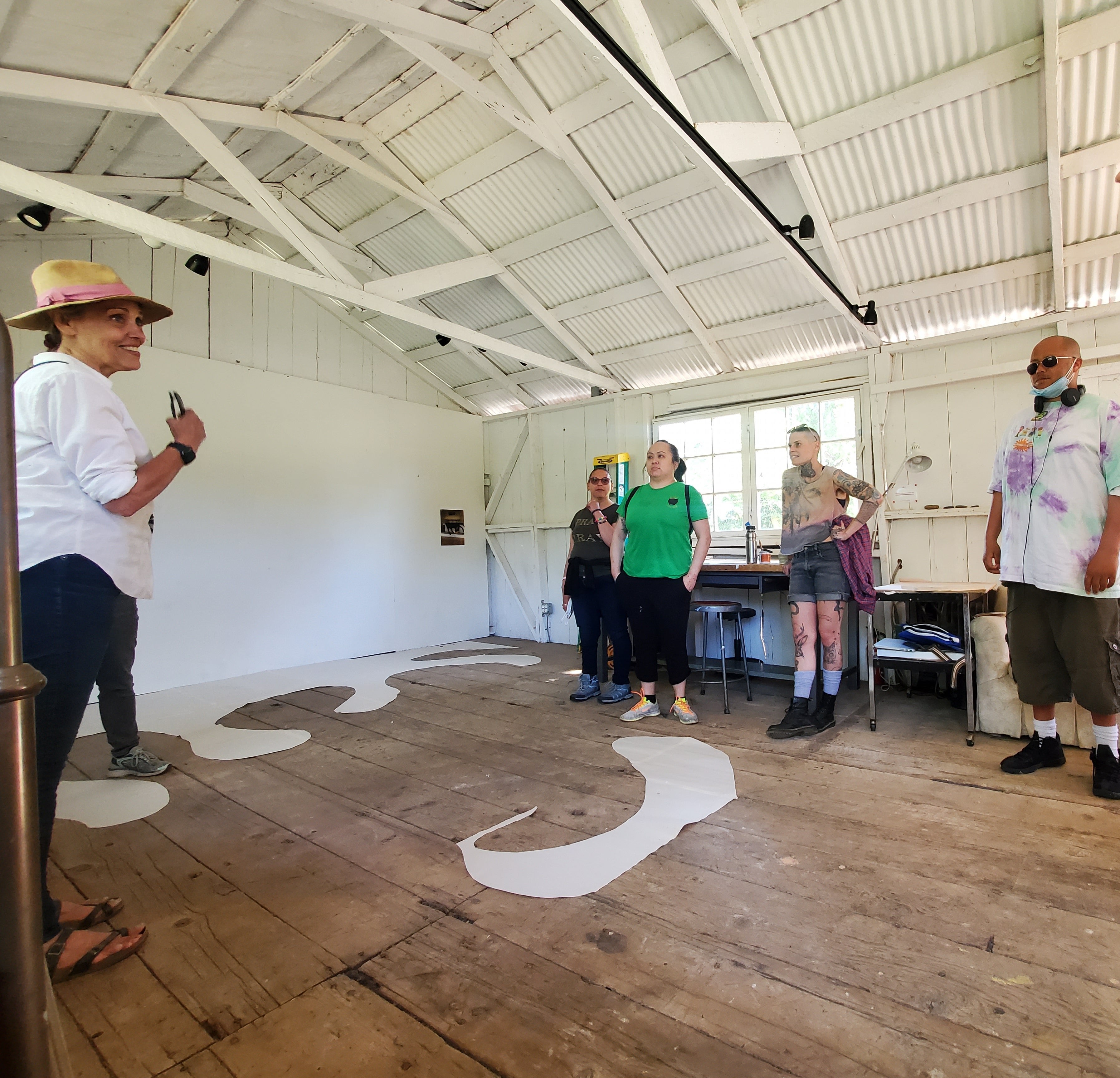 five people standing in a converted barn with rough wood floors and white vaulted ceilings. Round wave-like scraps of paper form an installation on the floor.