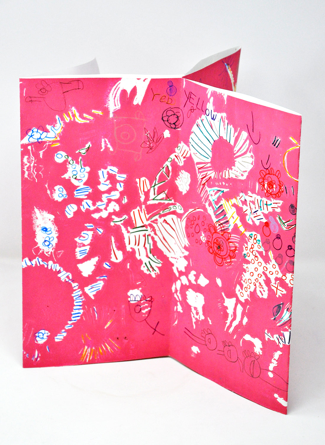 a handmade accordion fold artist book photographed standing on a white backdrop. The spread of pages in focus are colored bright pink, and have white, blue, and red symbols drawn over their entire surface. There are also several words referring to colors handwritten and integrated into the design. 