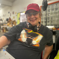 A portrait photograph of a person in an art studio. The person is a woman with light skin and glasses. She is smiling and looking at the camera. She wears a red baseball cap, pink headphones around her neck, and a black tee shirt with an image of an orange truck with headlights ablaze. There background is blurred, but it's clear there are large windows, tables, and artwork hanging on the walls. 