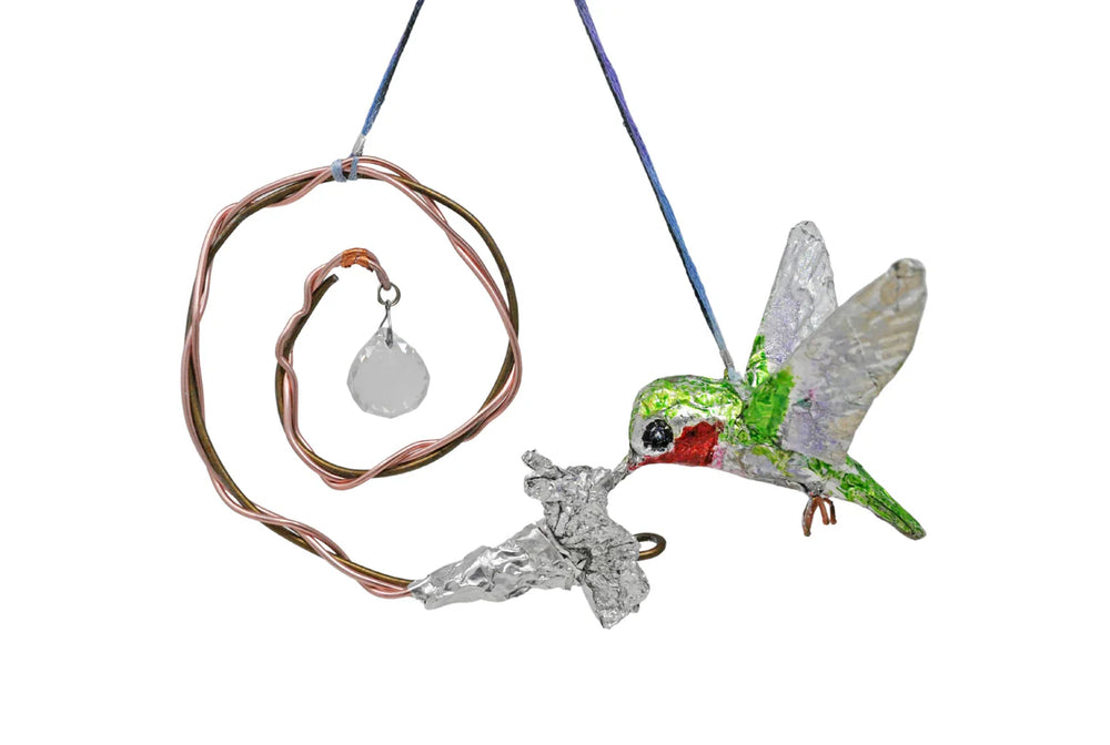 A hanging mobile sculpture in the shape of a realistic hummingbird drinking nectar from a flower. The object is made using various materials, including wire, foil, tape, beads, and other recyclable media. In this image, the hummingbird is positioned on the right side. It has a green and white body and a red spot under its chin. Its wings are flung wide as if it's hovering in mid-flight. Its beak is inserted into a silver foil flower. The flower has a copper spiral stem growing from a diamond bead.