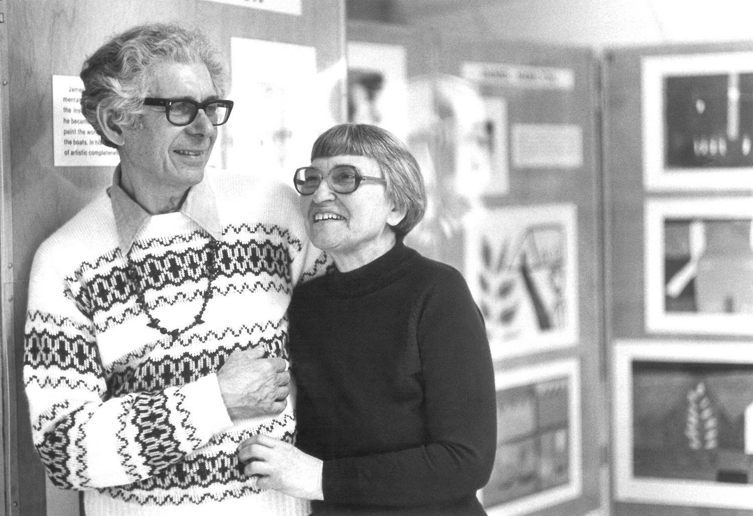 A grainy black and white photograph of two people standing in front of a gallery wall. The figure on the left has short wavy hair and wears dark rimmed glasses and a knit sweater. They have their arm around the other figure, who has short straight hair and is wearing glasses and a dark sweater. The wall behind them is hung with multiple prints and drawings. 