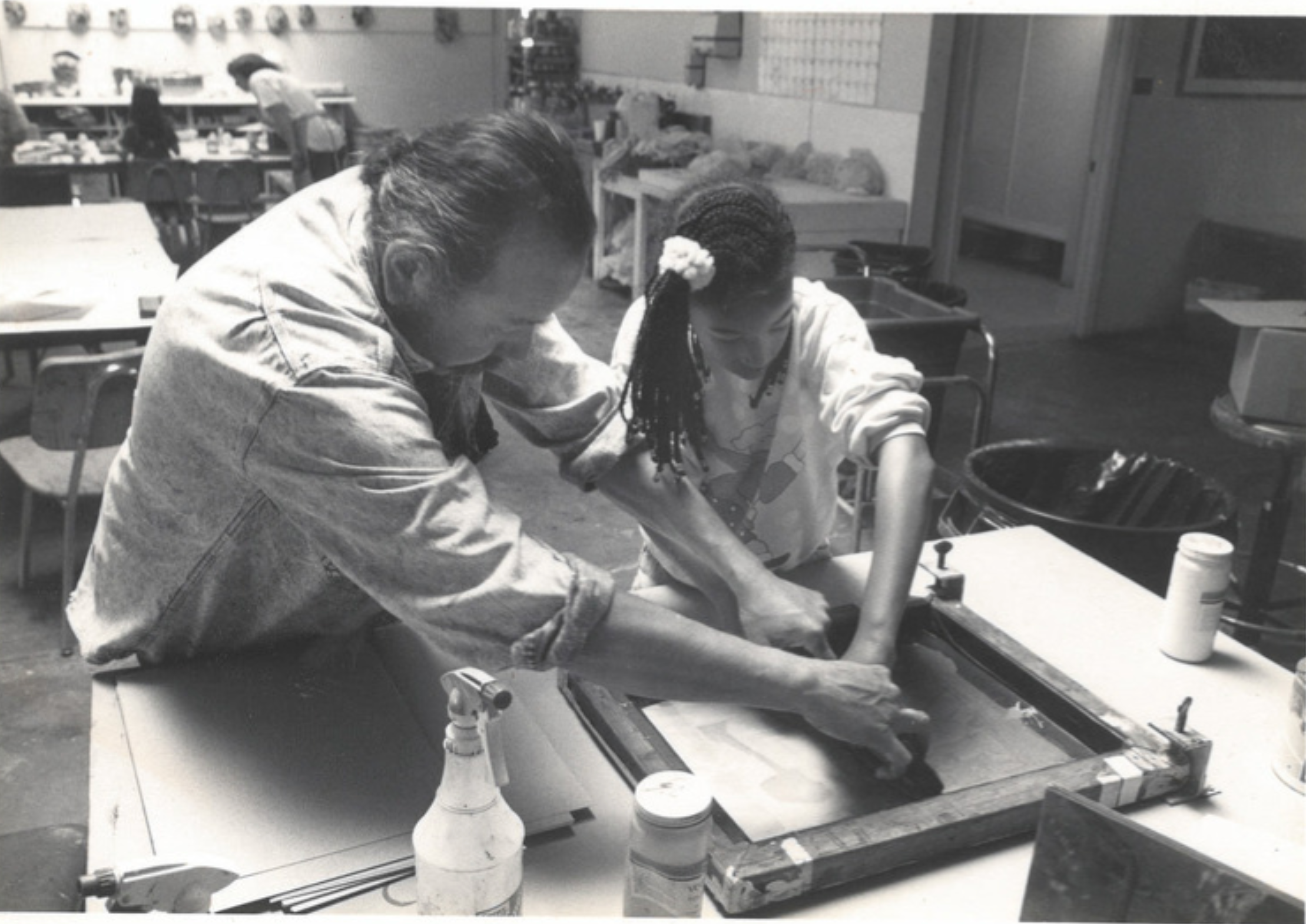 A grainy black and white photograph of two people working in a printmaking studio. The figure on the left has long hair pulled into a pony tail and a mustache. The figure on the right has long braided hair in a pony tail and wears a light sweatshirt. They are working together to apply ink to a screen. There is a stack of paper, ink containers, and a spray bottle on the table in front of them. Behind them, there are more work tables and art hanging on the walls.  