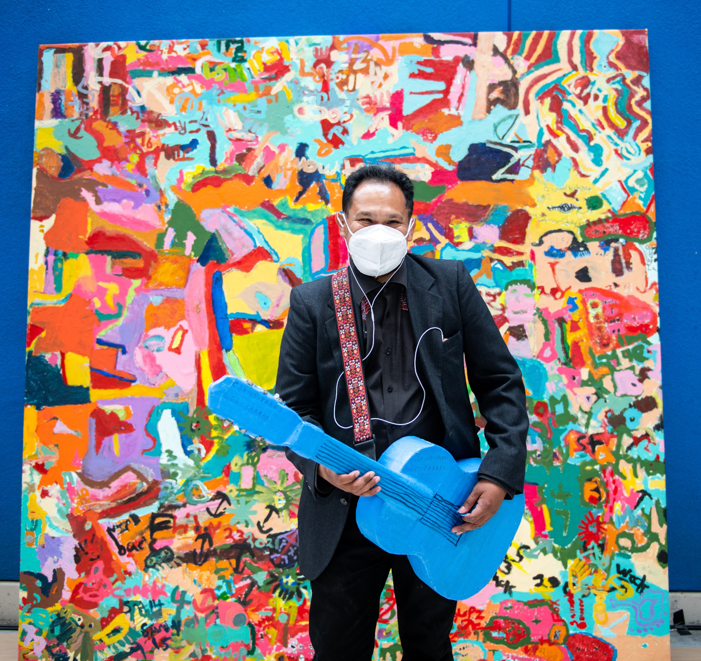 a photograph of a person standing in front of a large colorful abstract painting. The person is a man with brown skin and black hair, wearing headphones, a face mask, and a black suit. He is holding a blue sculpture shaped like a guitar. 