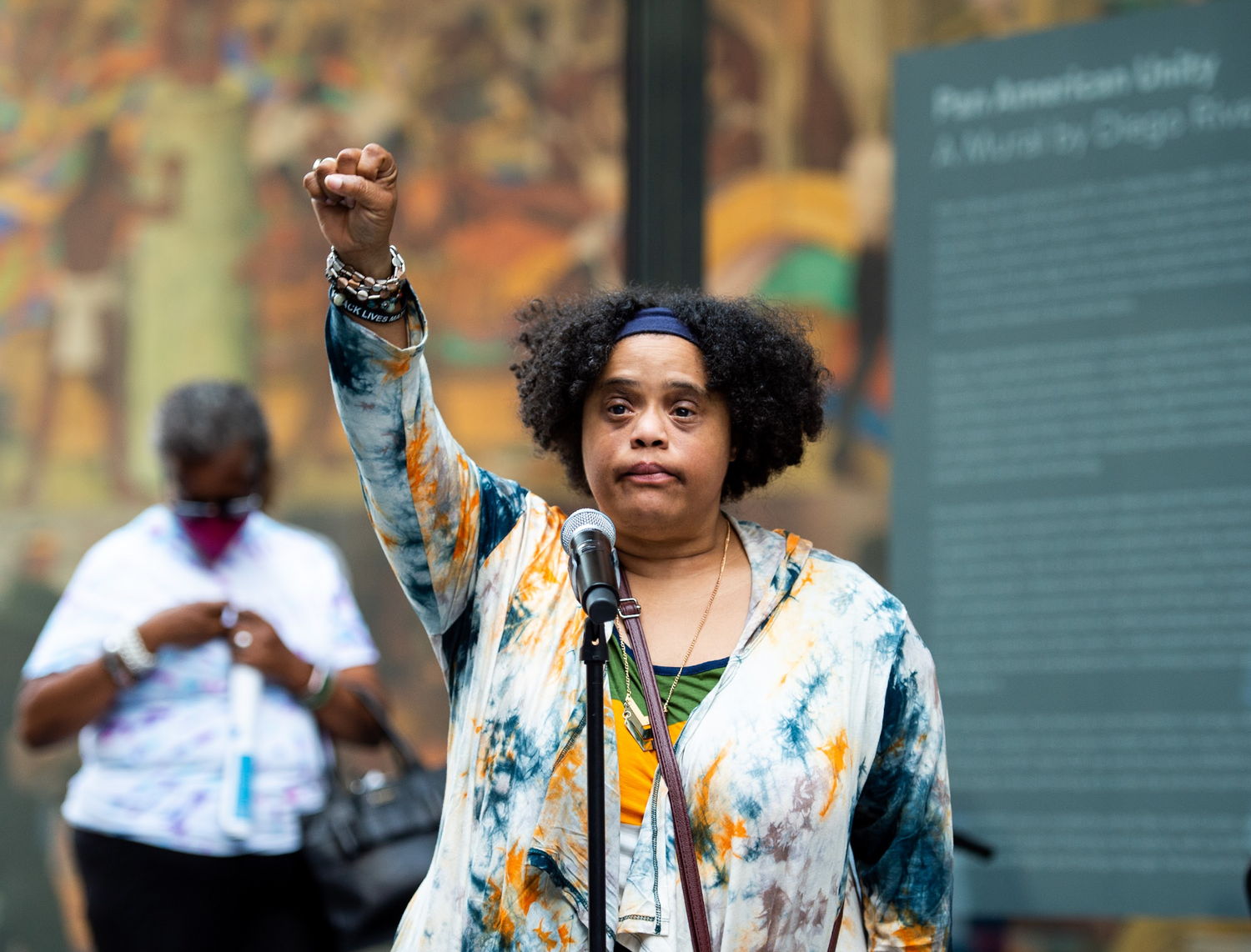 a photograph of a person standing outside in a museum courtyard, before a microphone on a stand. The person is the artist Halisi Noel-Johnson, a black woman with short curly black hair, a tie-dyed sweatshirt and a blue headband. She raises her fist in a powerful gesture, and wears a bracelet that reads "black lives matter." She is photographed having just finished addressing the crowd. The background is blurred but there appears to be a large green sign, and another figure wearing a mask and a white shirt.