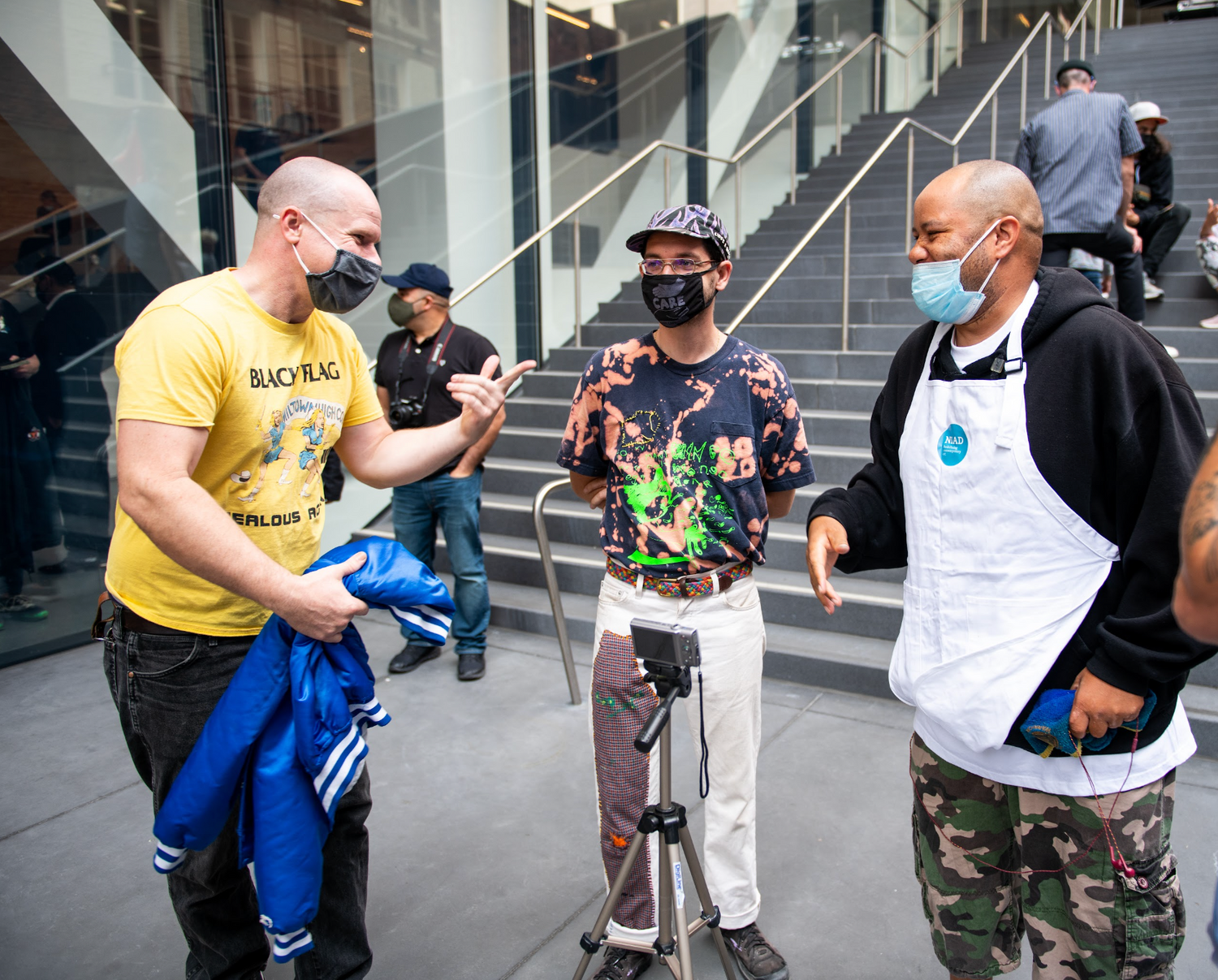 A photograph of several people talking in a museum courtyard. The figure on the left wears a yellow teeshirt, jeans, and a dark face mask. They gesture toward the person on the right, who is laughing and wearing a mask, a black sweatshirt, a white apron, and camo pants. There is a camera on a tripod set up between them, and a person with a baseball cap standing nearby. Figures mill around in the background on a tall stairway that leads into the museum.