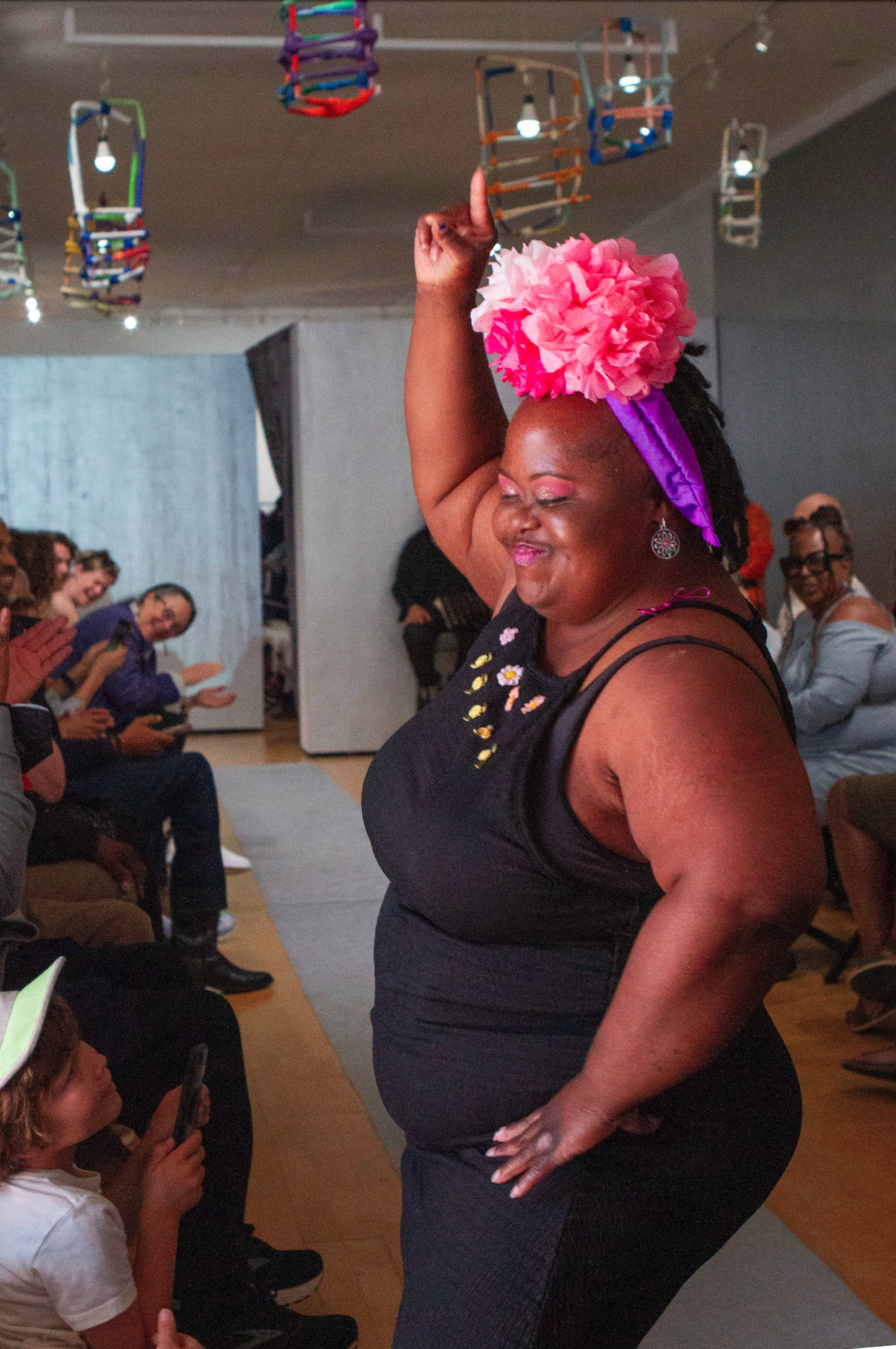 A photograph of a person walking down a fashion runway. The model is artist Nakiesha White, a black woman wearing a large pink paper flower on her head and a sleeveless black dress. She is pointing toward the ceiling with one hand and has her other hand on her hip. In the background, a crowd of people on either side of the catwalk applaud. The walls are painted silver and there are colorful paper lanterns installed on the ceiling.