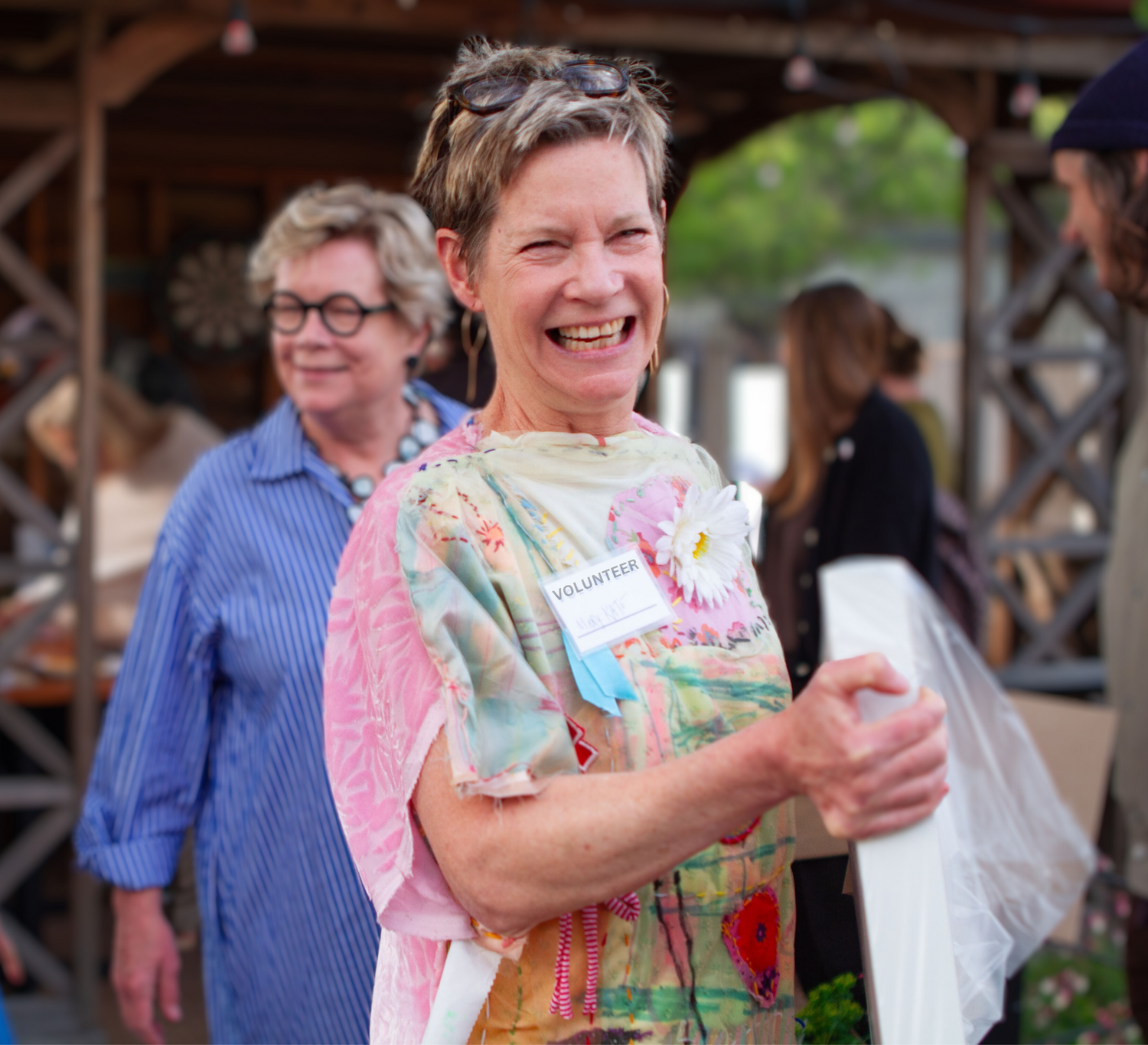 A photograph of several people standing outside. The figure most in focus is in the foreground . They have short blond hair, light skin, and a hand-sewn dress made of pink and green fabrics with a white fabric flower sewn near the neckline. The person is smiling at the camera, carrying a wrapped canvas, and wearing a badge that says "Volunteer." In the blurry background, there are other people milling around and looking at art on tables.