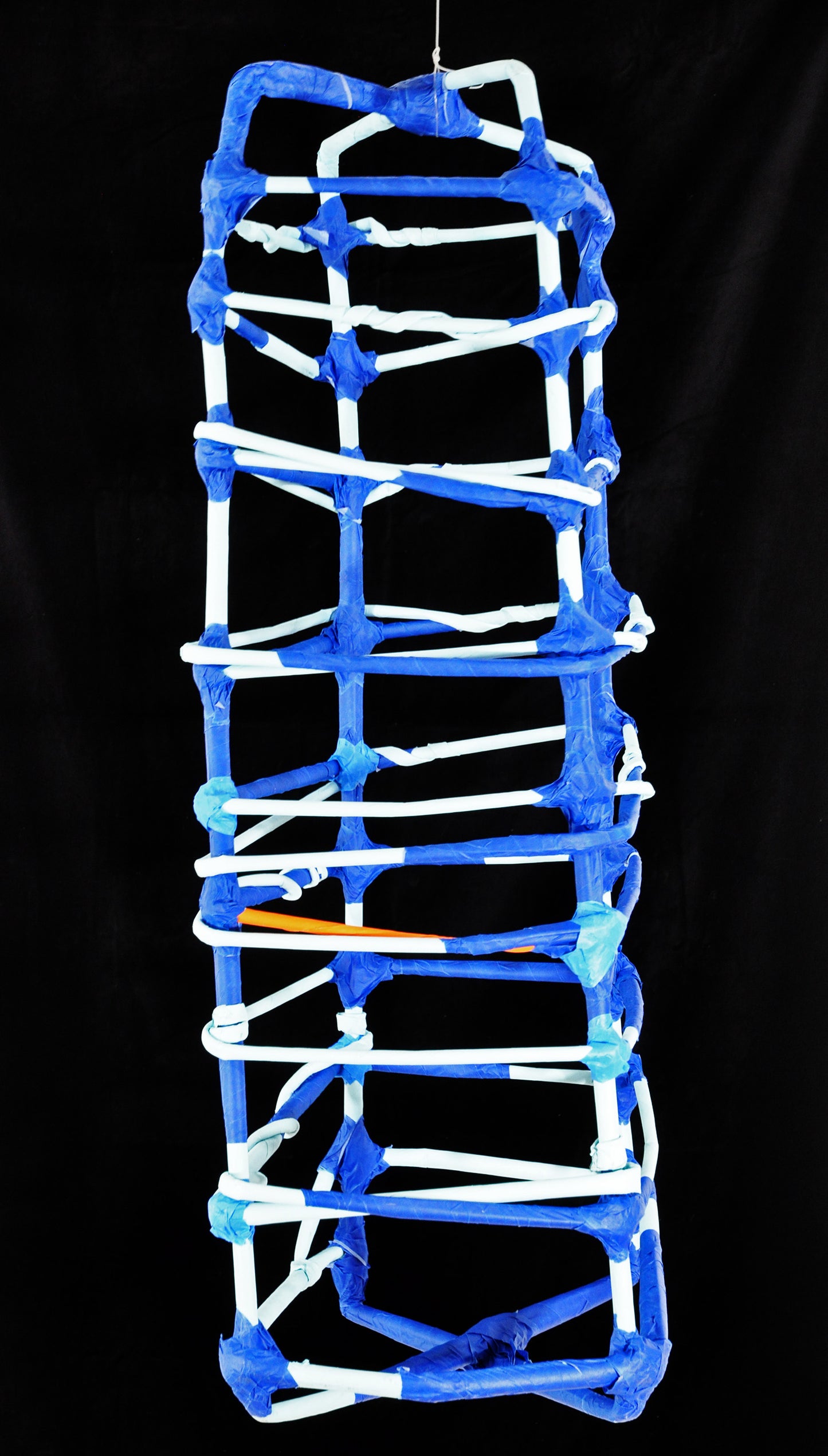 a sculpture made of tightly rolled tubes of paper and blue painters tape, photographed against a black backdrop. The form is a dimensional grid or cage-like structure. The white paper tubes are bound by blue tape wherever they intersect. 